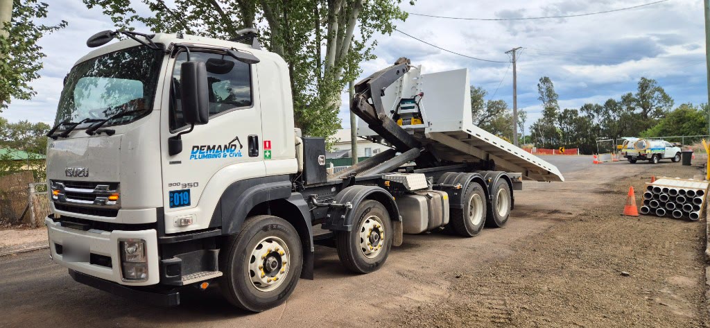 White Isuzu hook-lift truck with a tilted flat-top tray and winch on a job site, with a Demand Plumbing & Civil support ute in the background.